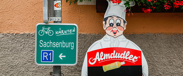 Cartello verde che indica Sachsenber, simpatico cartonato pubblicitario Almdudler, cascata di fiori rosa