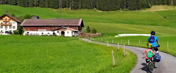Famiglia in bicicletta lungo un percorso panoramico della Ciclabile dei Tauri, circondata da prati verdi e montagne.