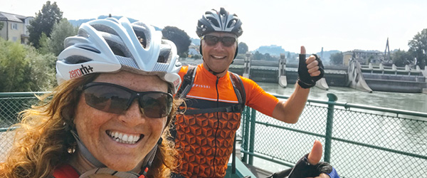 Coppia in bicicletta con casco e occhiali da sole lungo un fiume in Austria.