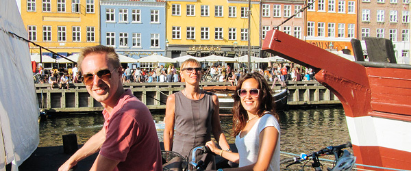 Turisti in bicicletta lungo Nyhavn a Copenaghen, con barche e case colorate sullo sfondo.