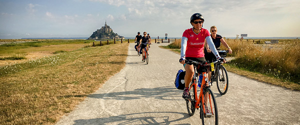 Ciclisti in vacanza con Girolibero su una strada panoramica verso Mont Saint-Michel, circondati da un paesaggio naturale.