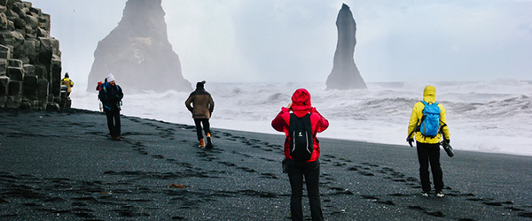 Gruppo di escursionisti in viaggio con Girolibero con k-way in una spiaggia nera camminano verso i faraglioni di Reynisdrangar, Islanda.