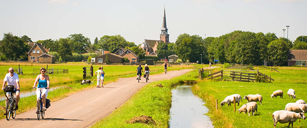 Ciclisti attraversano un piccolo villaggio con canale, mulino a vento e vegetazione rigogliosa sotto un cielo luminoso.