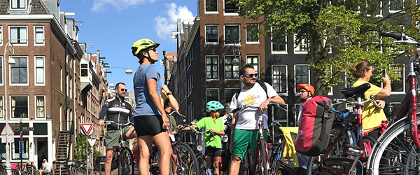 Gruppo di persone in bicicletta in viaggio con Girolibero ad Amsterdam, vicino a edifici tipici della città. Viaggiare con Giroliboer, in famiglia.