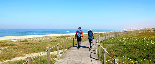 Coppia che cammina su una passerella in legno tra le dune di una spiaggia nel nord del Portogallo, sotto un cielo limpido.