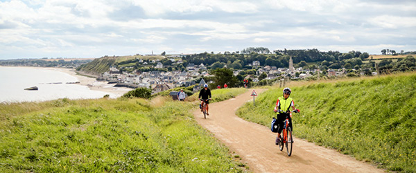 Cicloturisti sulla pista ciclabile lungo la costa della Normandia con vista sulla baia vicino a Arromanches-les-Bains, Francia