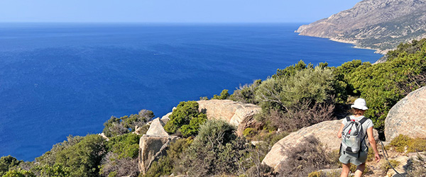 Escursionista lungo un sentiero panoramico tra grandi rocce granitiche e macchia mediterranea, con vista sul Mar Egeo, in Grecia.