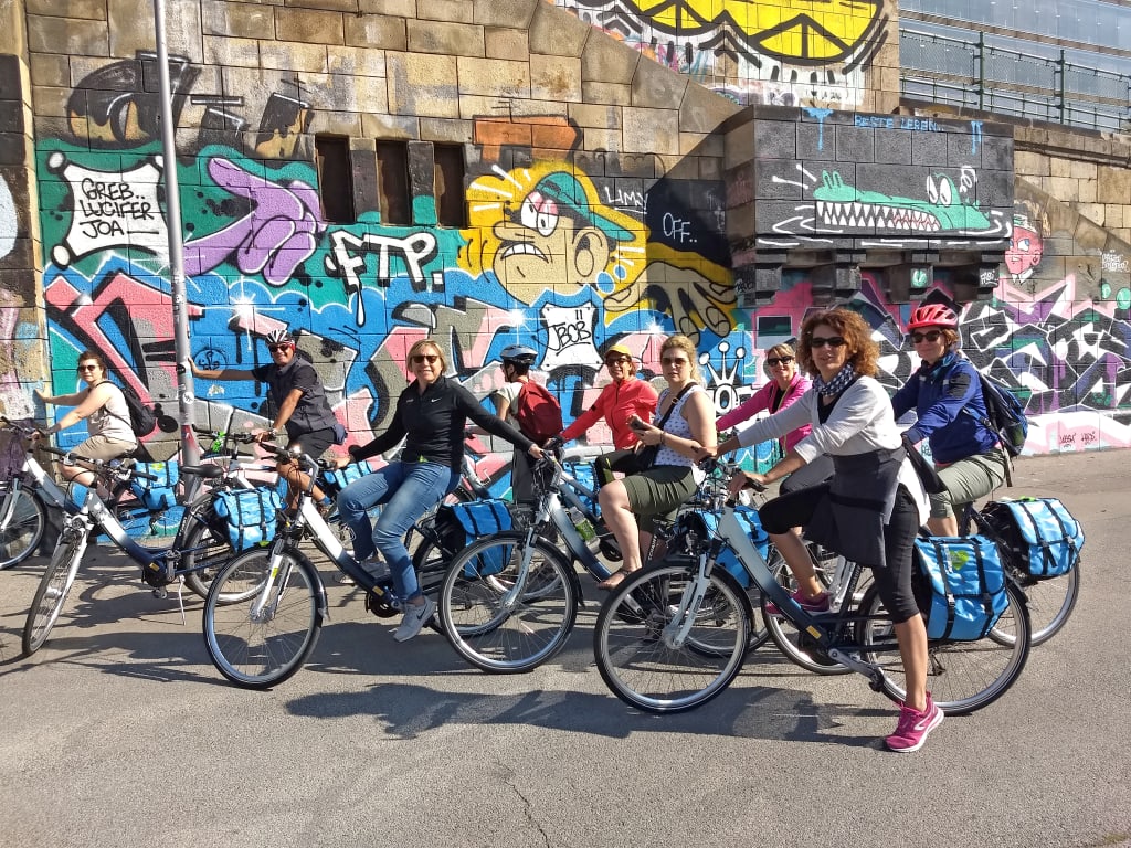 Gruppo di ciclisti in sosta davanti a murales urbani a Vienna, viaggio in bici tra arte e quartieri alternativi