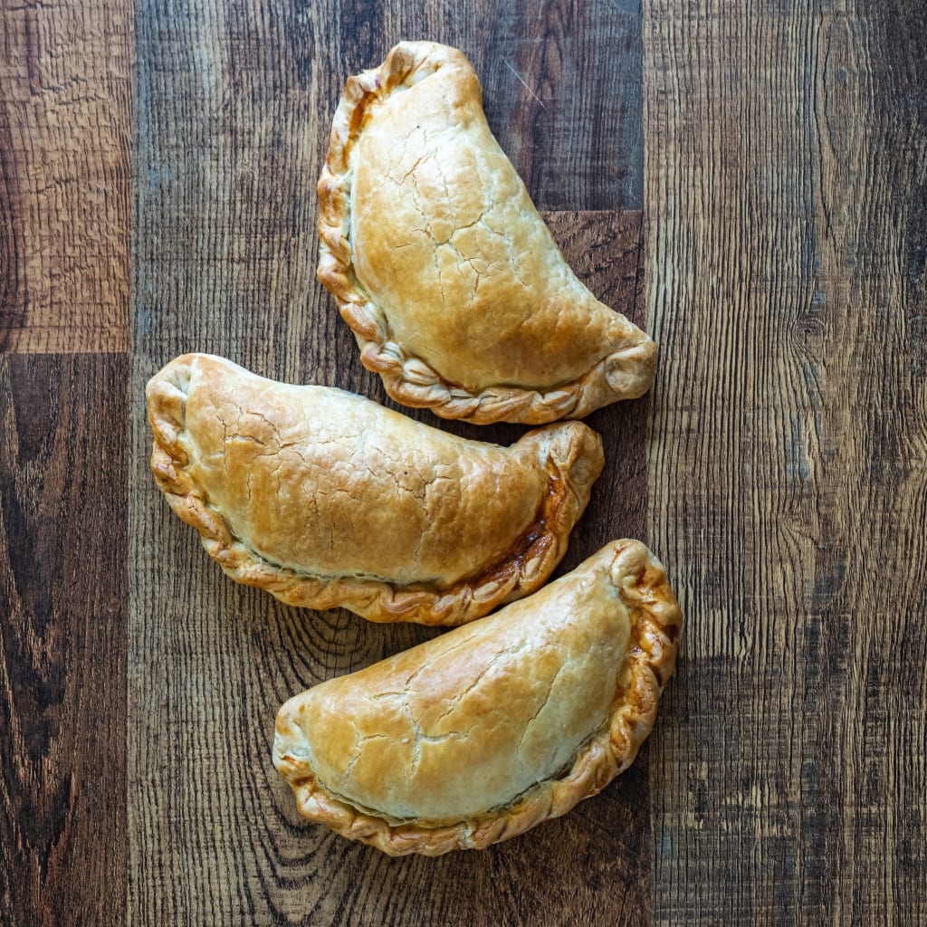 Cornish pasty su tavola in legno – Tre tradizionali pasticci della Cornovaglia (Cornish pasty) disposti su una tavola di legno, tipico street food locale.