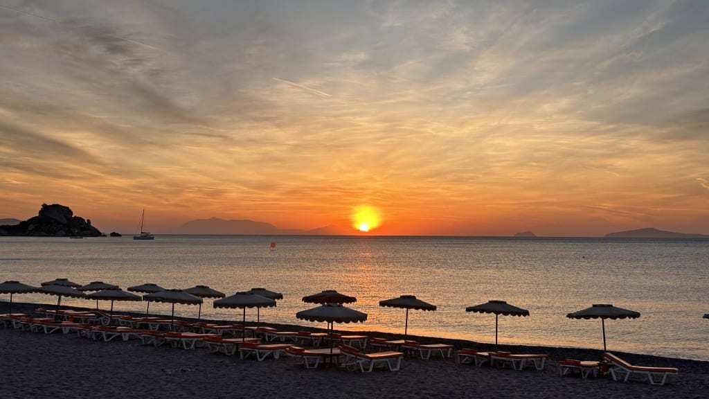 Tramonto su una spiaggia di Kos con ombrelloni e sfumature arancioni.