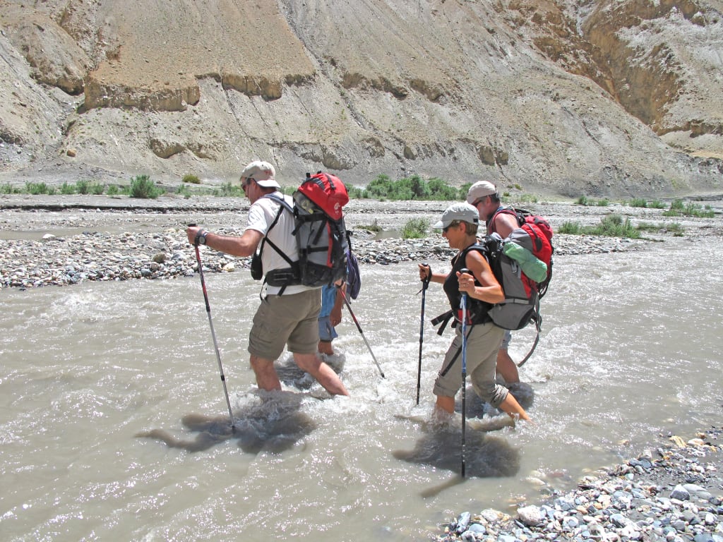 Escursionisti che attraversano un fiume con bastoni da trekking in un paesaggio montano del Ladakh.