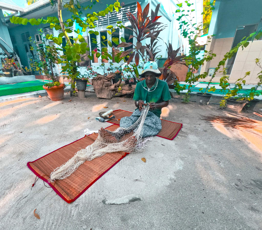 Pescatore maldiviano che ripara una rete da pesca davanti a una casa colorata su un’isola locale delle Maldive.