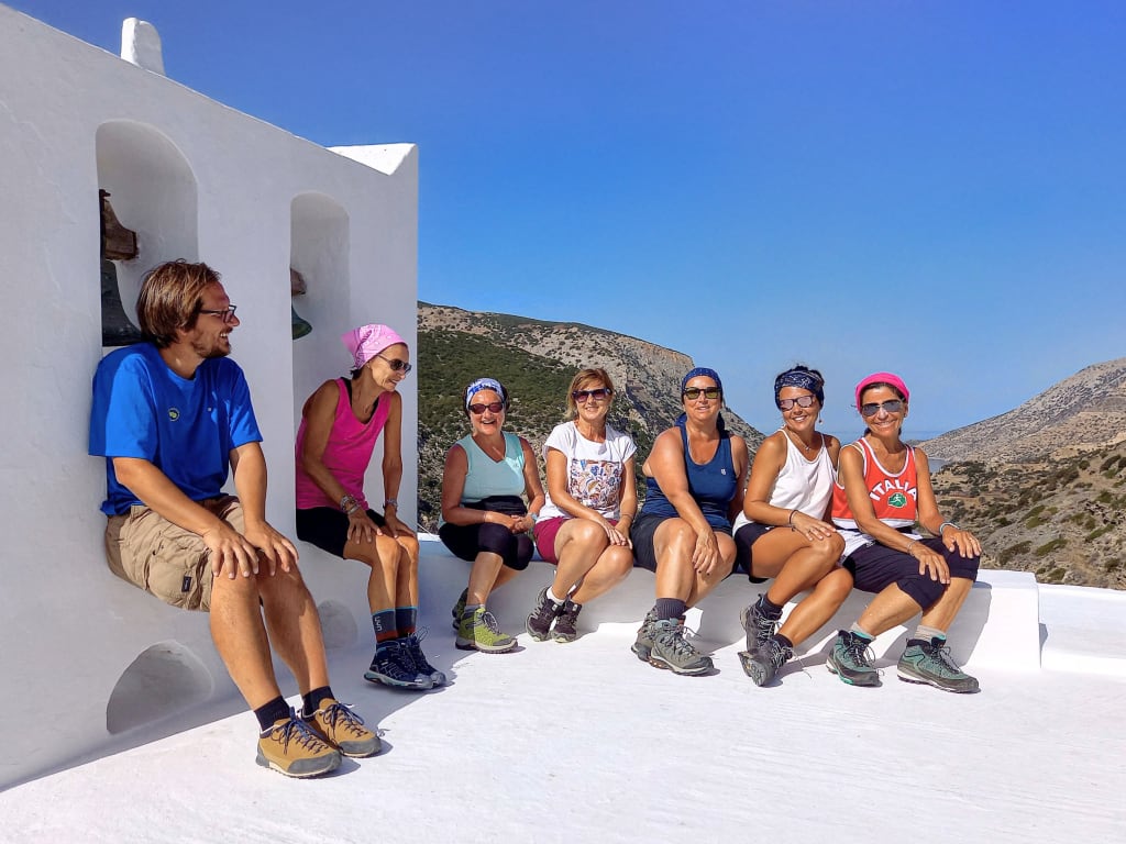 Gruppo di turisti in viaggio con Girolibero seduti sui gradini bianchi di una casa tipica in Grecia, con vista sul mare.