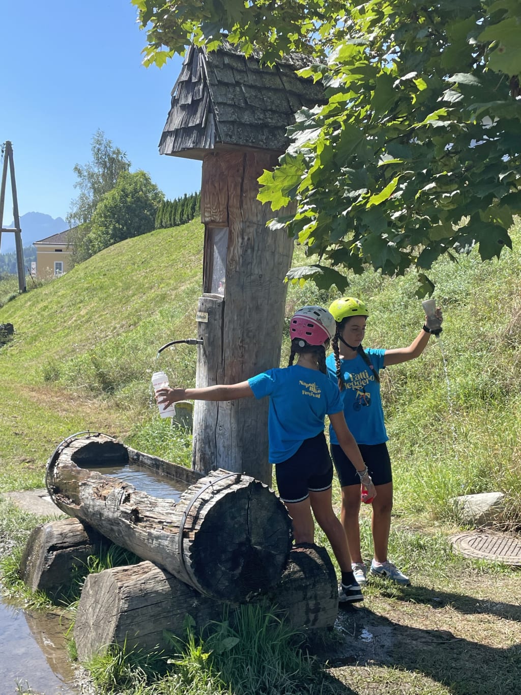 Ciclisti vicino a fontana in legno con vista sulle Alpi Giulie lungo la ciclovia Alpe Adria