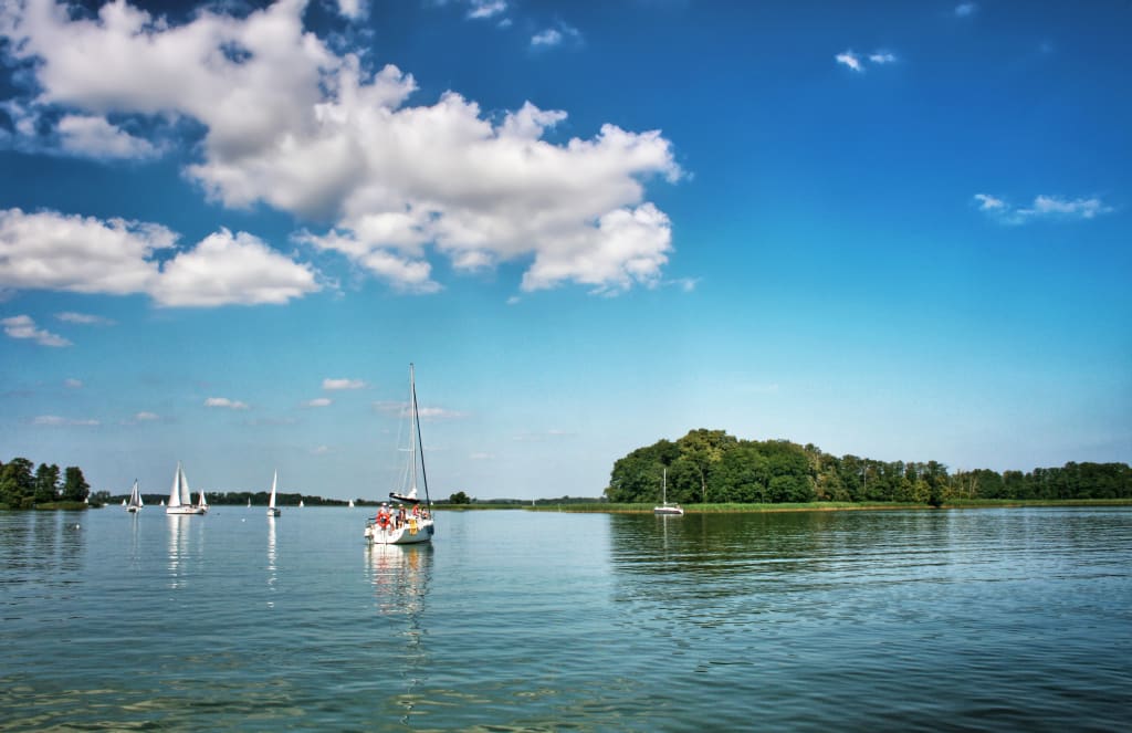 Barca a vela su un lago limpido nei Laghi Masuri, Polonia, con cielo azzurro e nuvole bianche.