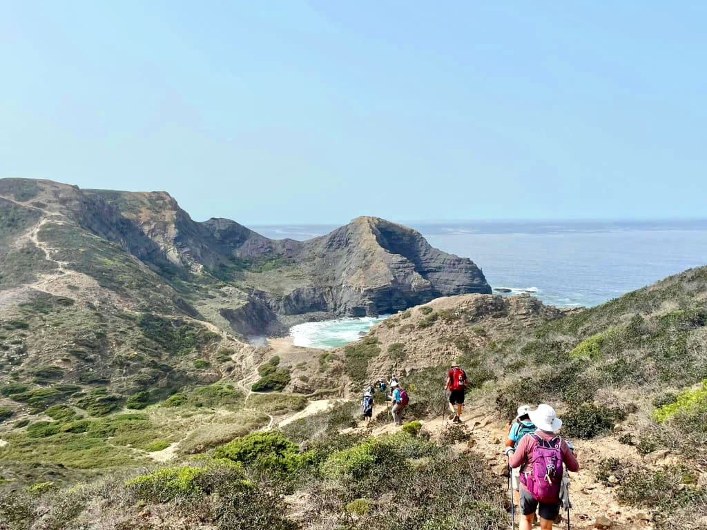 Escursionisti che percorrono un sentiero tra colline rocciose con vista sul mare, Rota Vicentina in Algarve