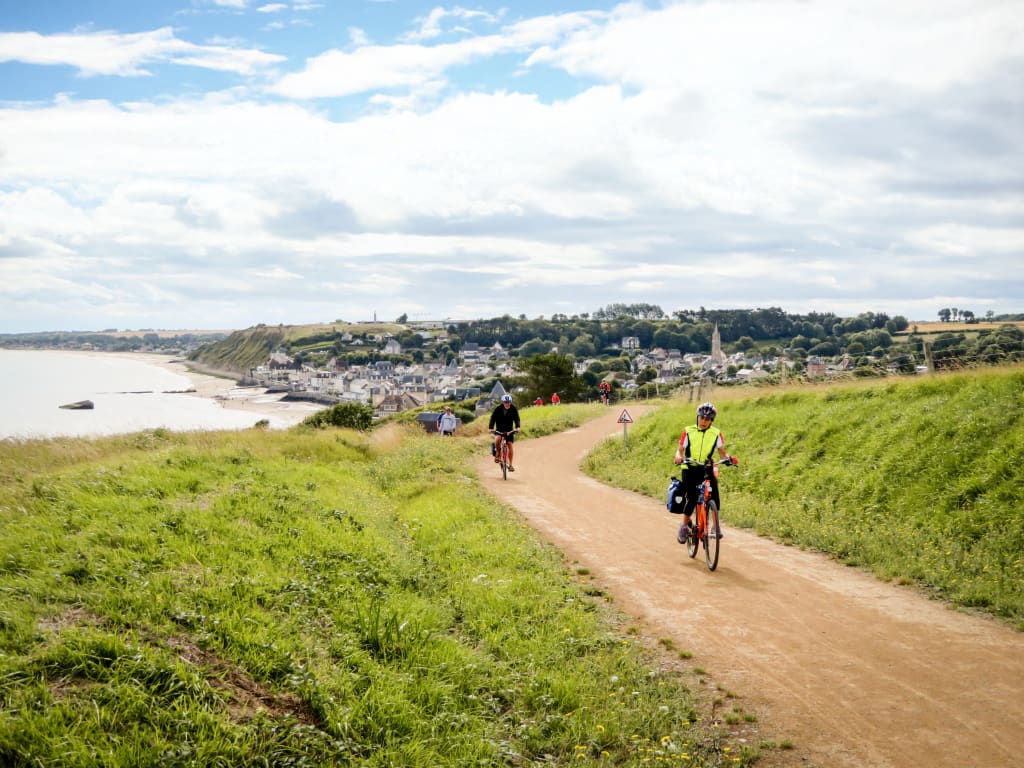 Cicloturisti sulla pista ciclabile lungo la costa della Normandia con vista sulla baia vicino a Arromanches-les-Bains, Francia