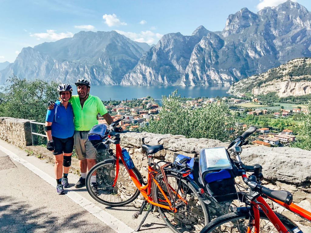 Coppia di cicloturisti in posa per una foto con lago e montagne, biciclette arancioni "Girolibero", ciclabile dell'Adige