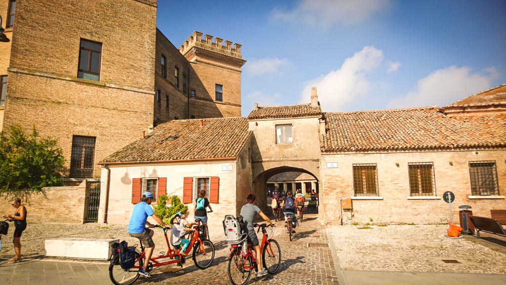 Famiglia in bici tandem passa davanti a una casa rurale nella campagna, viaggi in bici "Girolibero" per famiglie, Parco naturale delta del Po