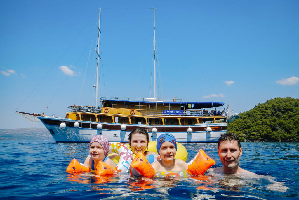 Family with children swimming in the calm waters of a bay in Croatia, with the boat "San Snova" behind them