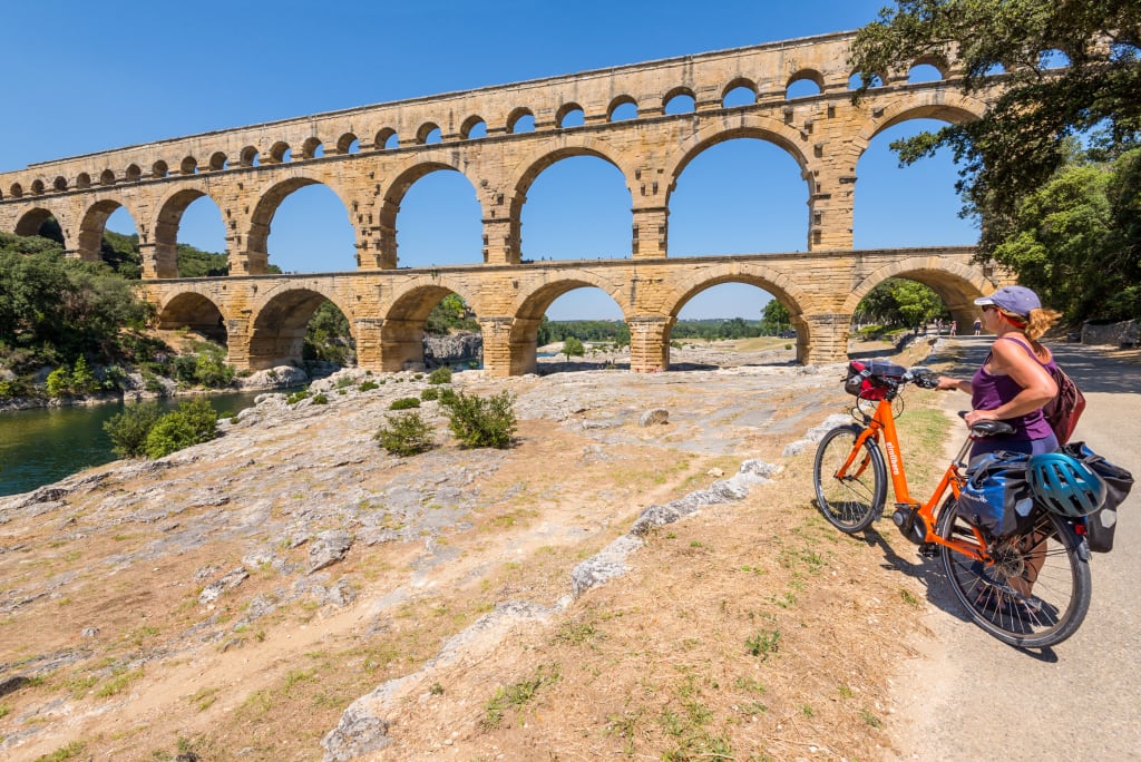 Cicloturista in viaggio con bicicletta arancione Girolibero, in posa davanti al maestoso ponte acquedotto romano Pont du Gard, Francia meridionale