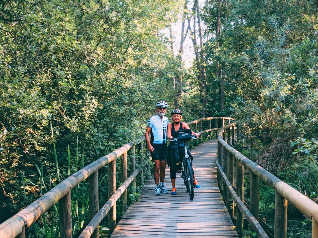 Coppia di ciclisti che attraversa una passerella in legno immersa nella natura, viaggi in bici "Girolibero"