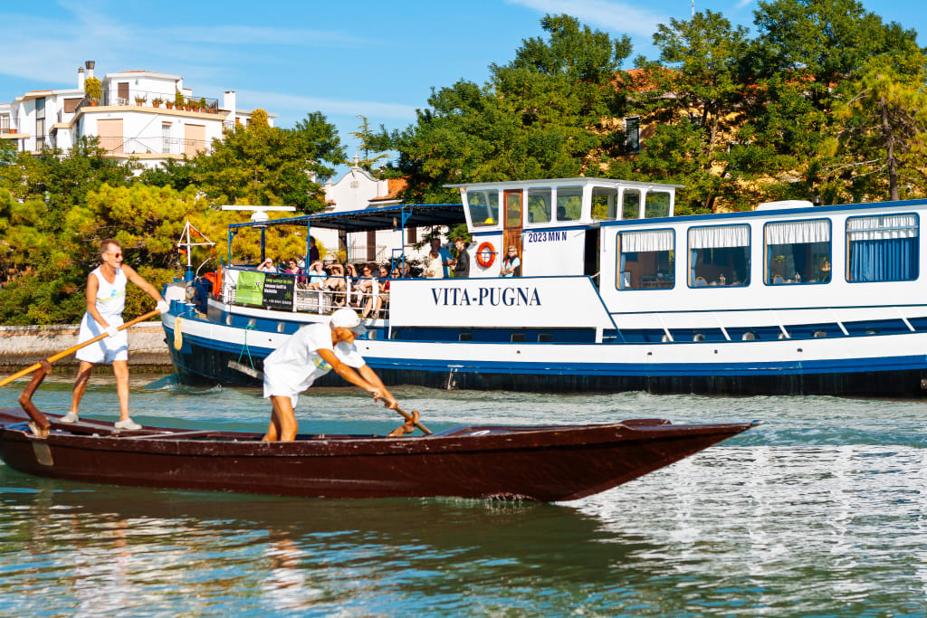 Due persone in barca remi in un canale con barca "Vita Pugna" a fianco
