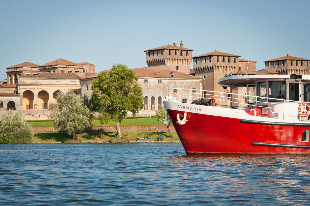 "Ave Maria“ in Mantua, Boot auf einem Fluss, mit historischen Gebäuden und Grünflächen im Hintergrund