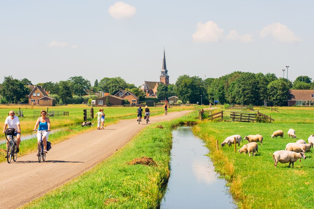 Ciclisti attraversano un piccolo villaggio con canale, mulino a vento e vegetazione rigogliosa sotto un cielo luminoso.