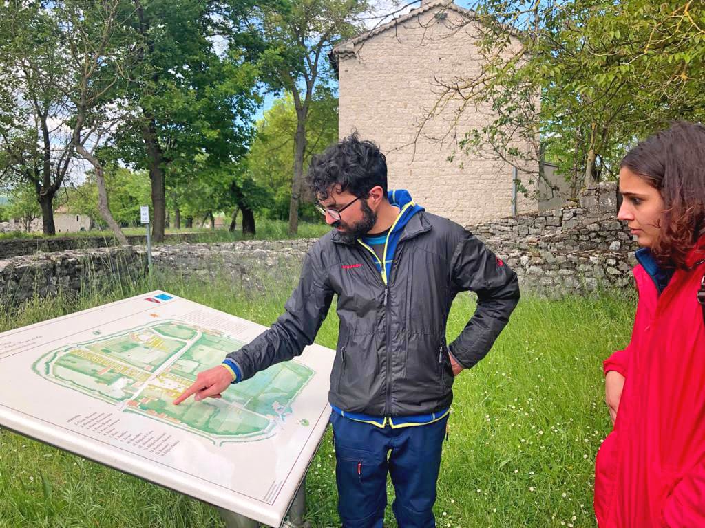 Guida ambientale che spiega una mappa storica a due viaggiatori in un sito all'aperto, vicino a un edificio antico.