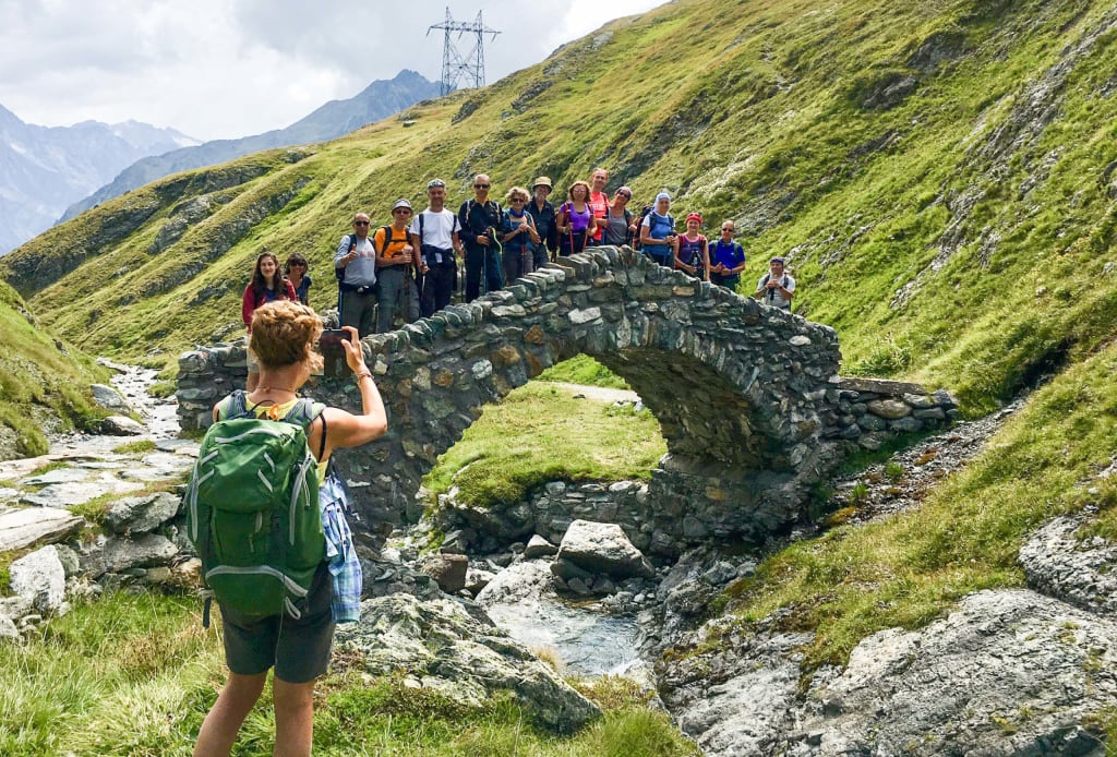 Gruppo di escursionisti fa una foto su un sentiero che attraversa un antico ponte ad arco in una valle verdeggiante.