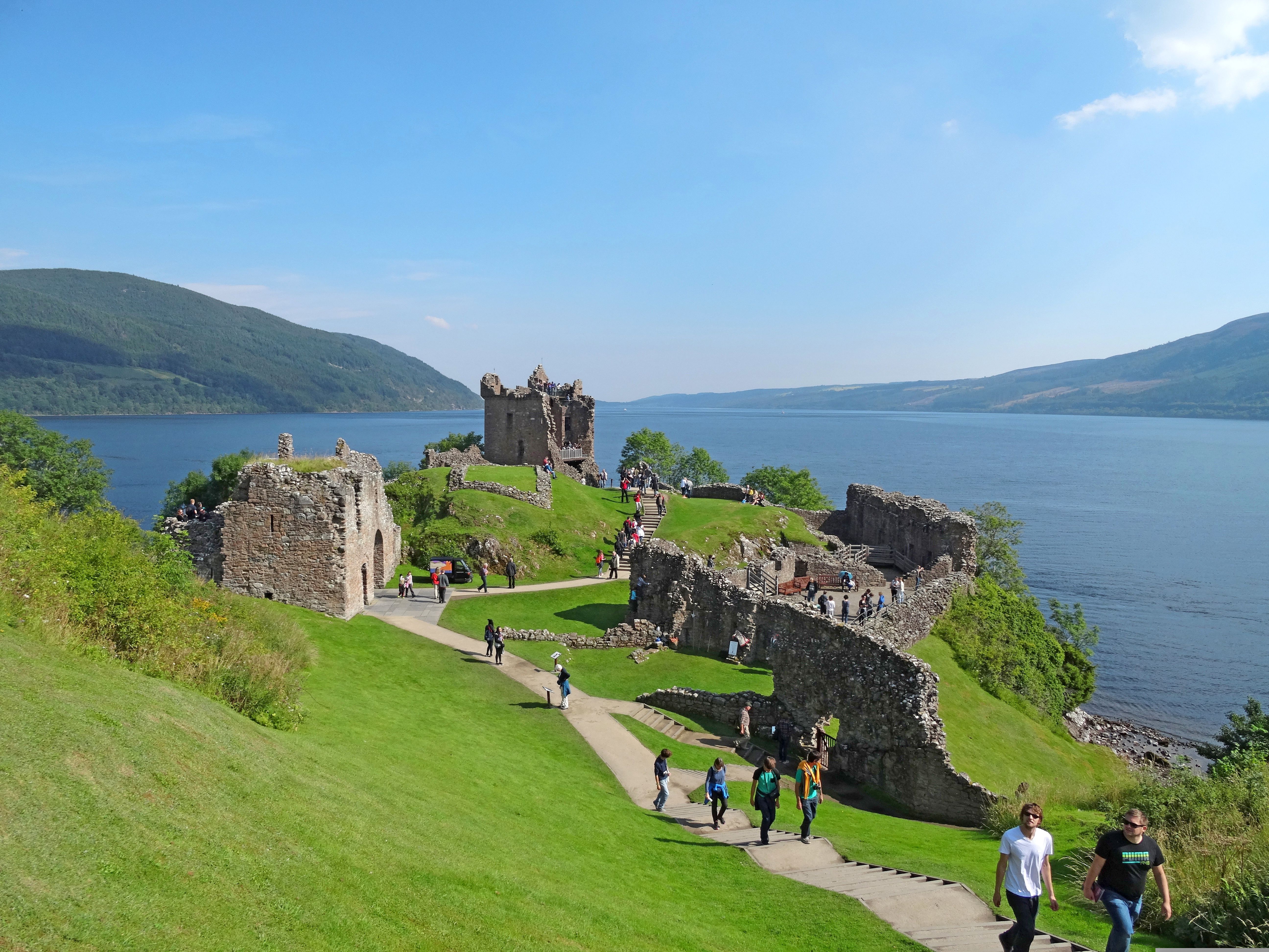 Veduta del castello di Urquhart sul lago di Loch Ness lungo il percorso del Great Glen Way, circondato da colline verdi e visitatori sul prato, Scozia.
