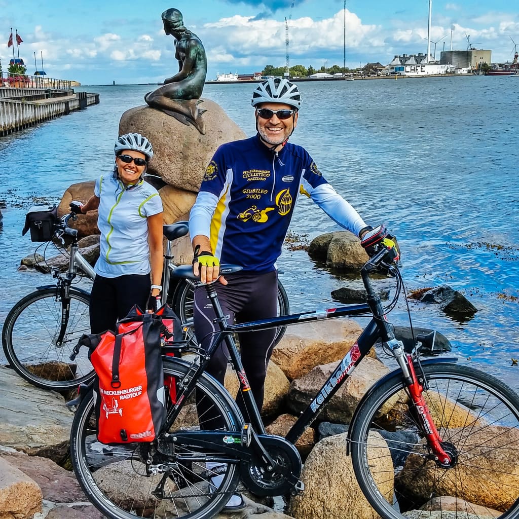 Coppia di cicloturisti davanti alla Sirenetta nel porto di Copenaghen, classica foto ricordo di un viaggio in bicicletta.