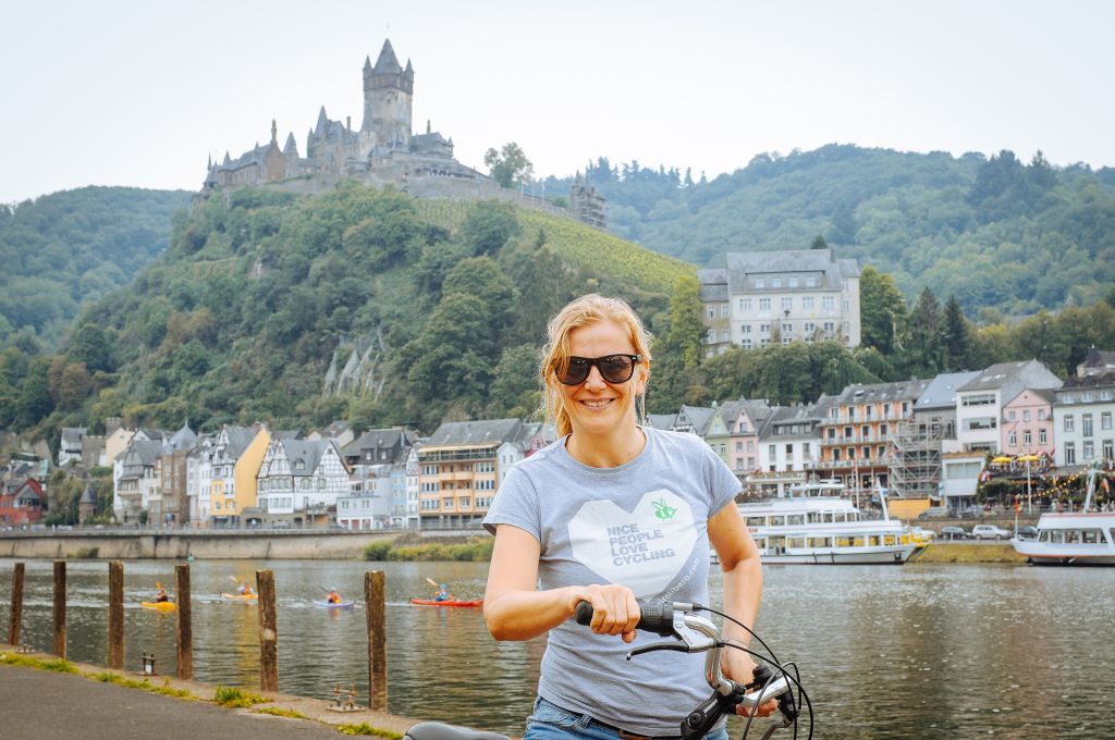 Cicloturista sorridente e con occhiali da sole lungo ciclabile della Mosella, castello imperiale di Cochem, Germania