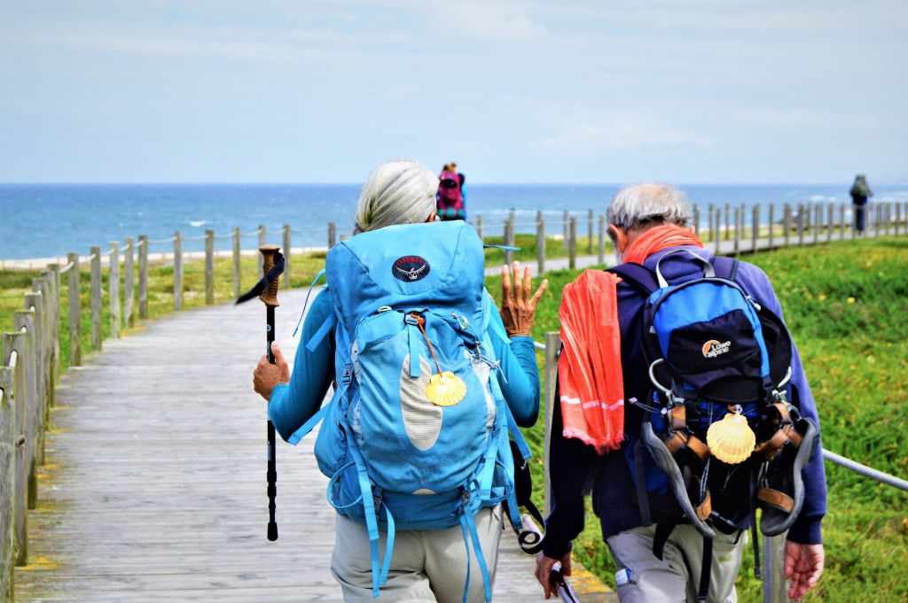 Due pellegrini con zaini che camminano su una passerella vicino alla spiaggia.