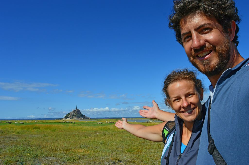 Coppia sorridente davanti a un paesaggio costiero della Normandia con vista su Mont-Saint-Michel, durante una giornata soleggiata.