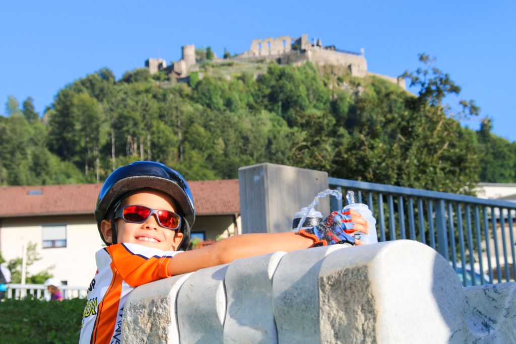 Bambino sorridente con caschetto intento a riempire la borraccia da una fontanella, ciclabile della Drava