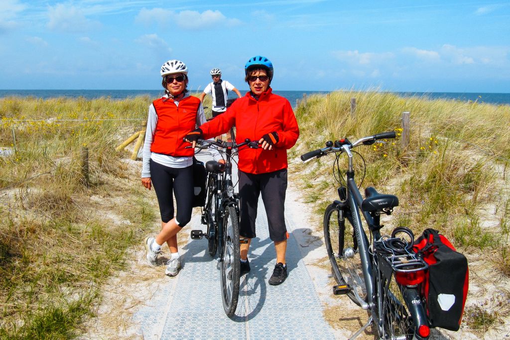 Ciclisti in viaggio con "Girolibero" fermi su un percorso ciclabile che attraversa dune sabbiose con vegetazione costiera lungo il Mar Baltico.