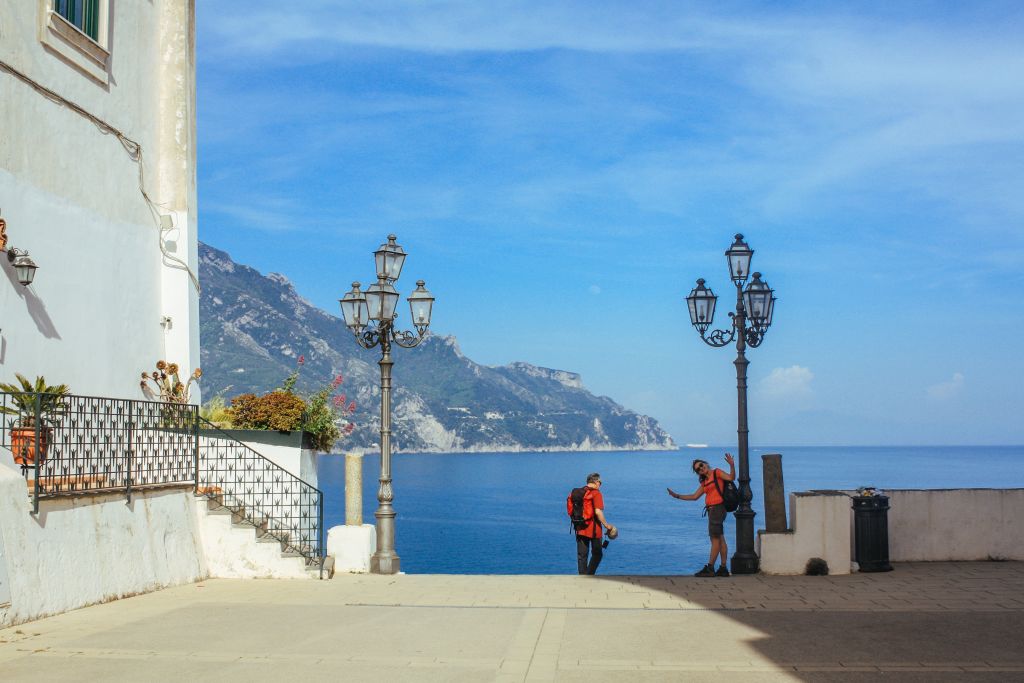 Escursionista sul lato un edificio bianco con piante e scalini con corrimano, mare limpido sullo sfondo, Costiera Amalfitana