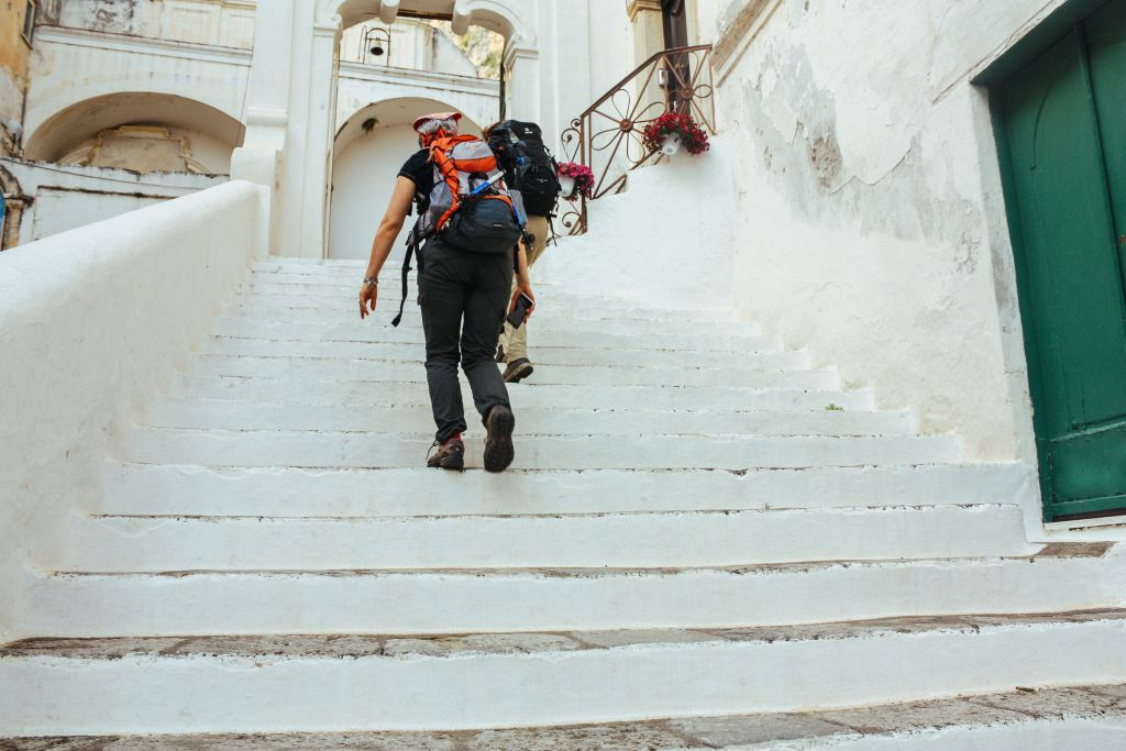 Coppia di escursionisti intenti a salire scale bianche, edifici bianchi, vasi floreaili, trekking, Costiera Amalfitana