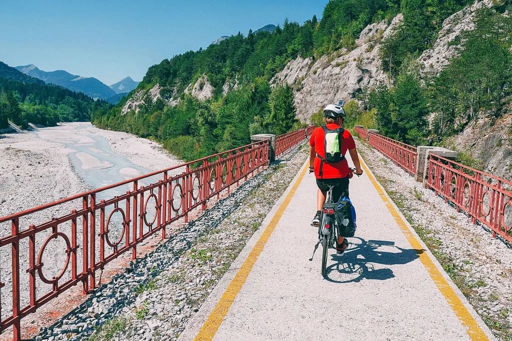 Cicloturista, pista ciclabie segnalata, ponte con ringhiere rosse, fiumicciatolo, paesaggio roccioso, Alpe Adria