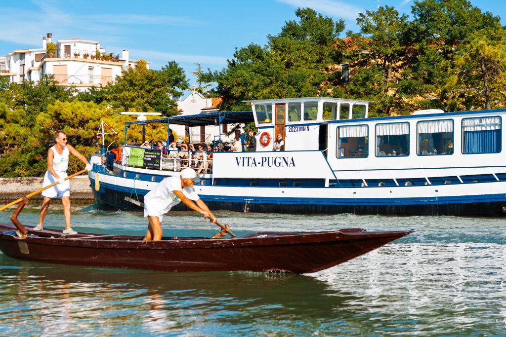 Due persone in barca remi in un canale con barca "Vita Pugna" a fianco