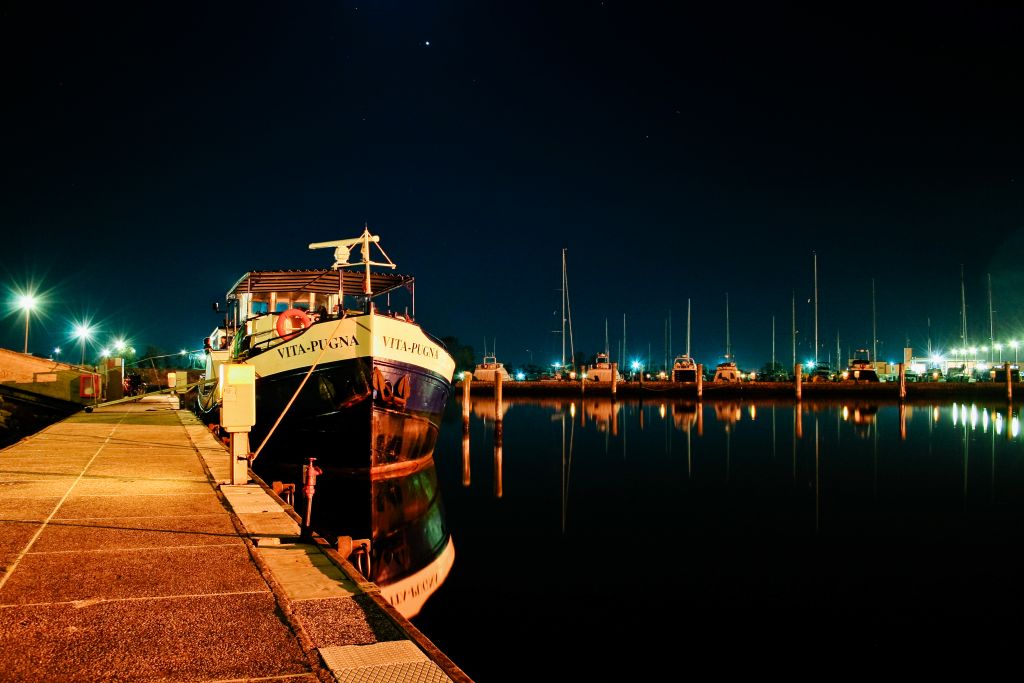 Barca "Vita Pugna" ormeggiata nel porto di Mantova di notte, con luci riflettenti sull'acqua.