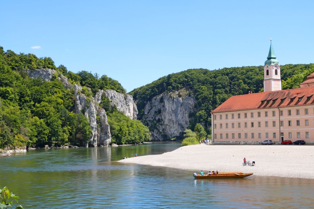 Monastero di Weltenburg lungo il fiume Altmühl, con spiaggia sabbiosa e canoe in riva all’acqua.