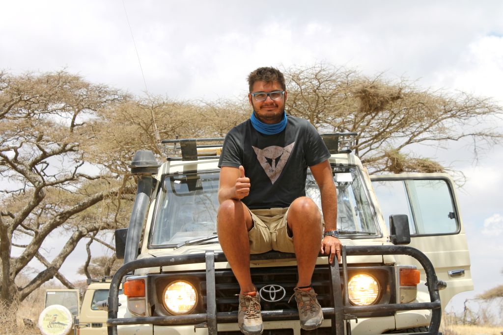 Persona seduta sul cofano di una jeep durante un safari, con alberi e savana sullo sfondo.