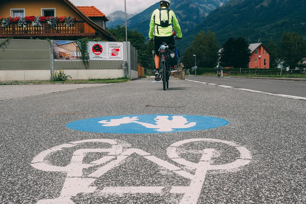 Cicloturista con caschetto, pista ciclabile segnalata, casa con balcone pieno di fiori, vacanza in bici, ciclovia Alpe Adria