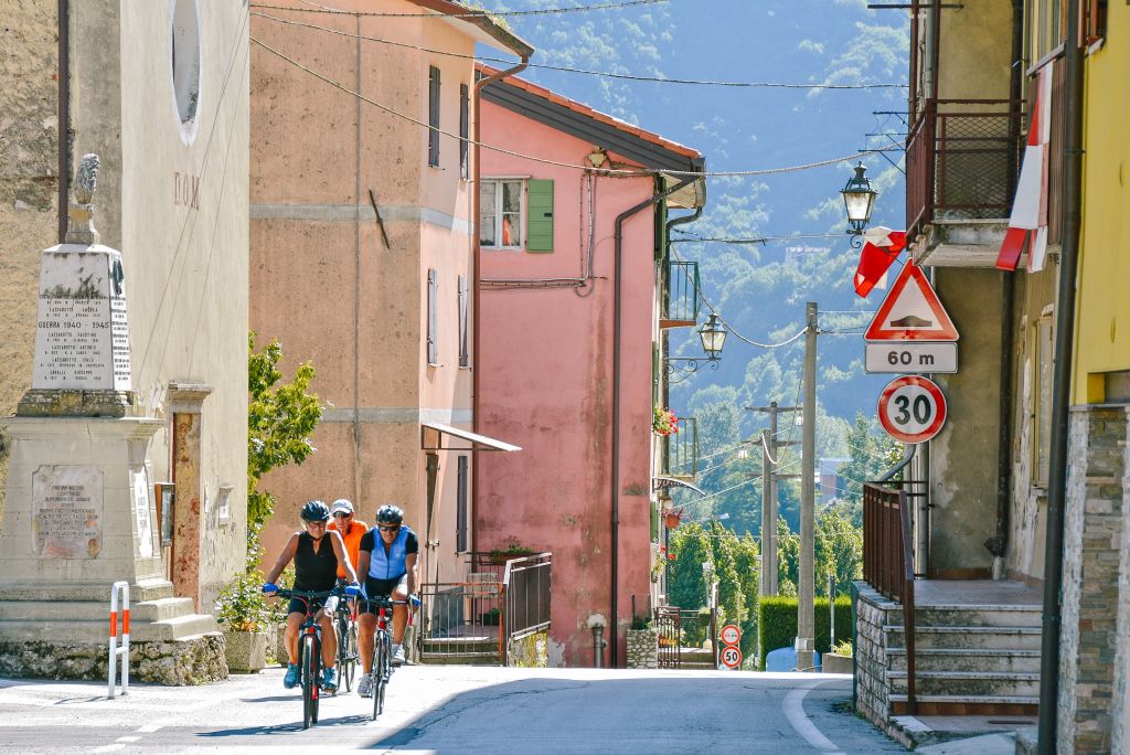 Ciclisti attraversano una stradina colorata con case tipiche nel centro storico di Bassano del Grappa, Veneto in bici