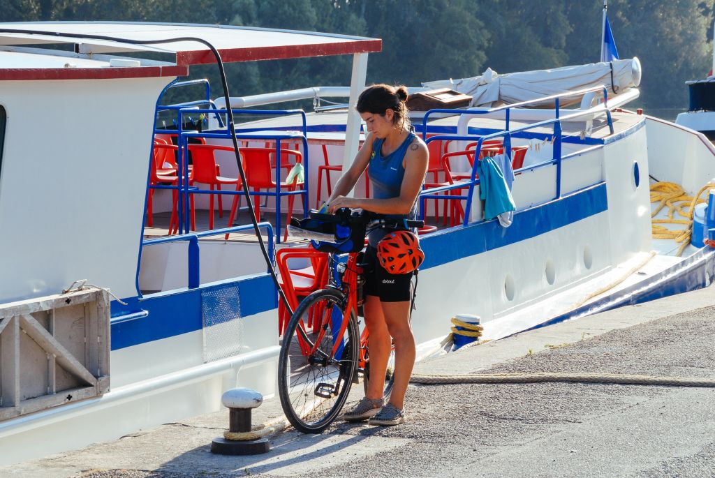 Ciclista con bici "Girolibero" davanti a una barca ormeggiata lungo fiume, Provenza.