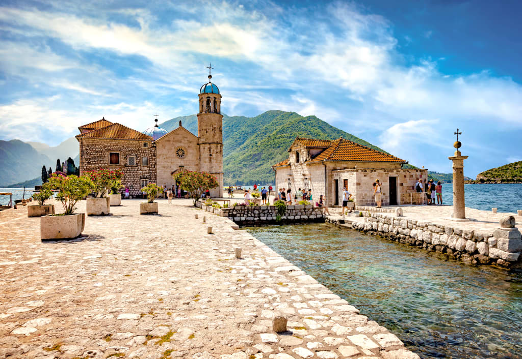 Chiesa storica e campanile con cupola azzurra, affiancata da un edificio storico, strada a ciottoli, acqua e colline verdi sulla baia di Kotor