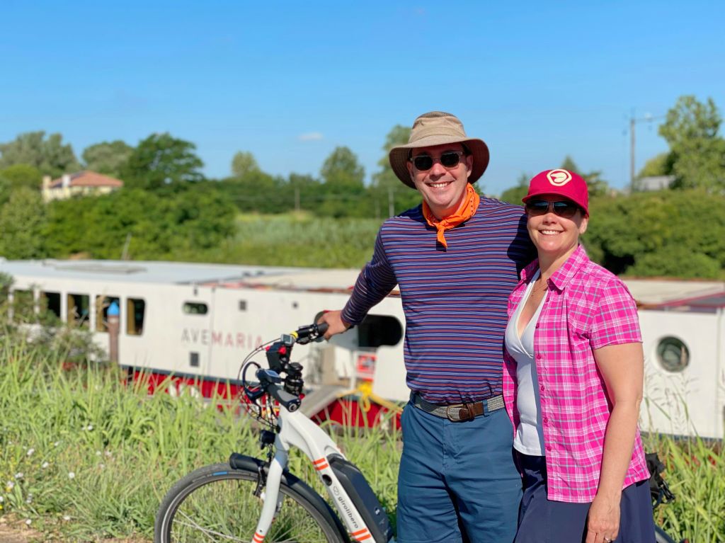 Vacanze in bicicletta, coppia sorridente fa una foto vicino ad un fiume, con chiatta Ave Maria ormeggiata sulle rive